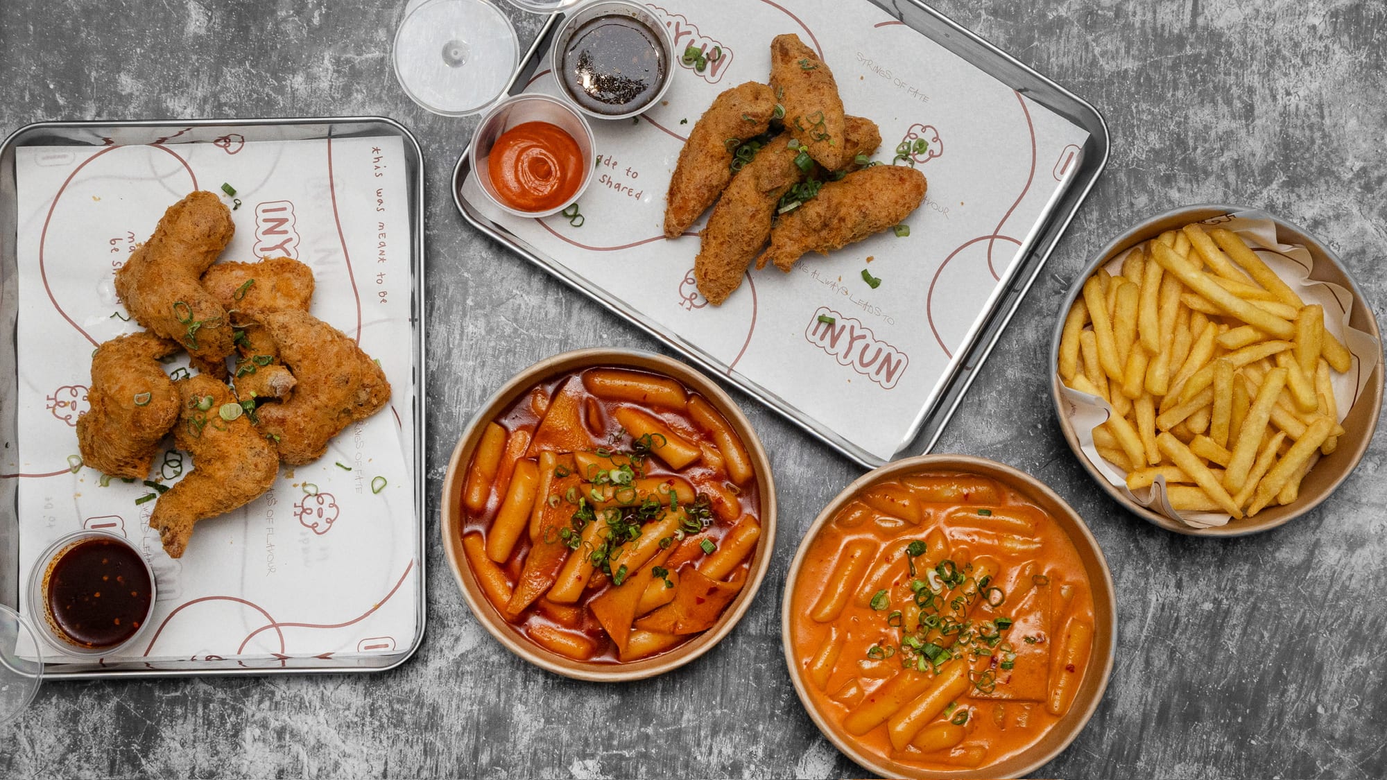 An overhead spread of INYUN Korean fried chicken with tteokbokki and chips on branded trays